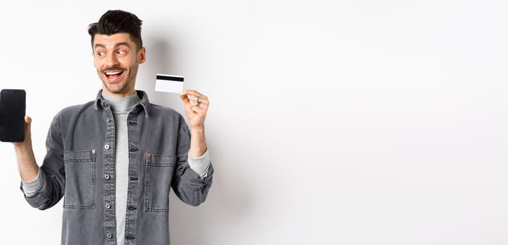 Handsome smiling man in denim jacket looking at empty smartphone screen and showing plastic credit card, good internet offer, standing against white background.