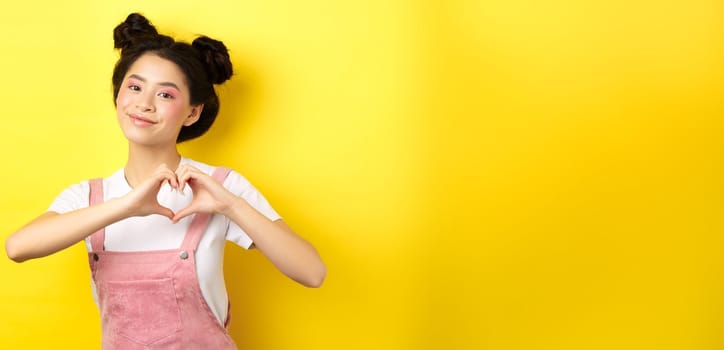 Valentines day. Cute asian girl sending her love, showing heart gesture and smiling at camera romantic, standing on yellow background.