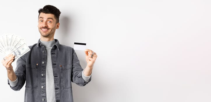 Happy guy showing plastic credit card and dollar bills, giving choice, standing on white background.