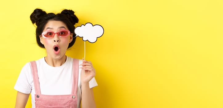 Summer and fashion concept. Excited party girl in sunglasses, holding comment cloud party mask and gasping amazed, standing impressed on yellow background.