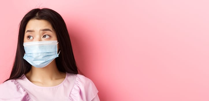 Covid-19, pandemic and quarantine concept. Close-up of sad asian girl in medical mask feeling lonely during coronavirus, standing against pink background.