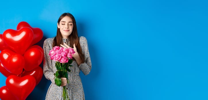 Valentines day. Romantic lovely woman in dress, close eyes and smiling, receiving flowers from lover, smelling bouquet of pink roses, blue background.
