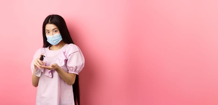 Covid-19, lifestyle and health concept. Beautiful asian woman in medical mask clean hands with sanitizer, apply antiseptic on palms, standing on pink background.