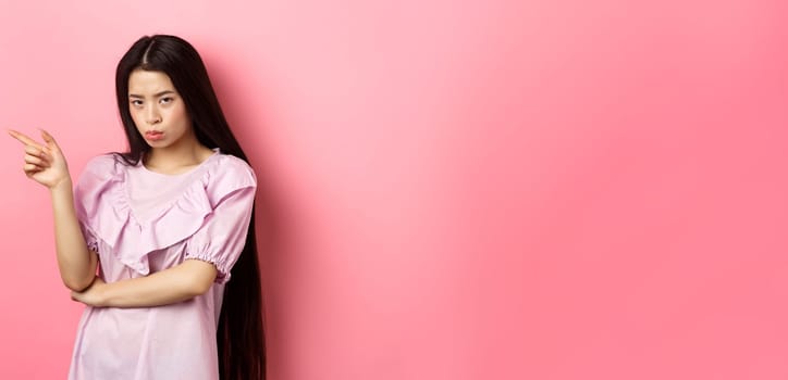 Sad and grumpy asian girl sulking, pointing left and look at camera jealous, want something, complaining and showing desired thing, standing on pink background.