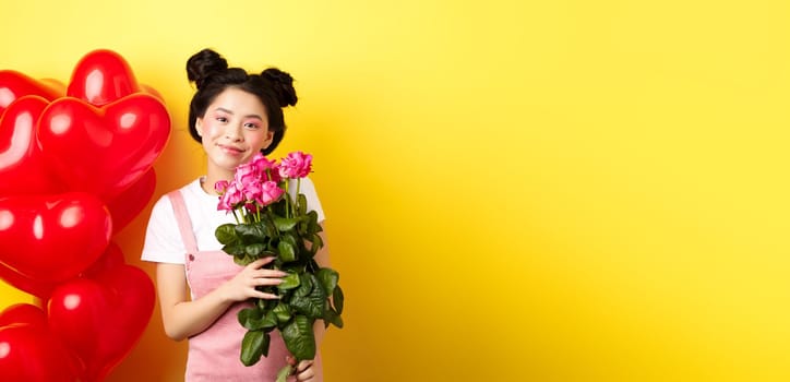 Happy Valentines day. Beautiful asian woman dressed for romantic date, holding bouquet of flowers and smiling. Girl with roses standing near heart balloons, yellow background.