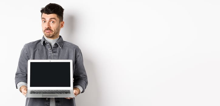 Indecisive young man pouting and shrugging while showing empty laptop screen, standing clueless on white background.