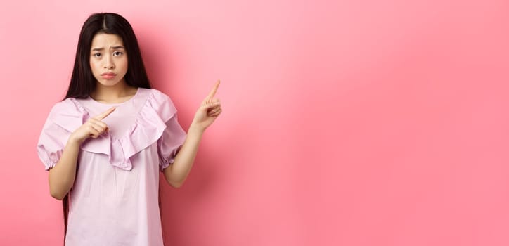 Sad and distressed asian girl frowning, sulking and pointing fingers right at disappointing bad news, feeling unhappy, standing against pink background.