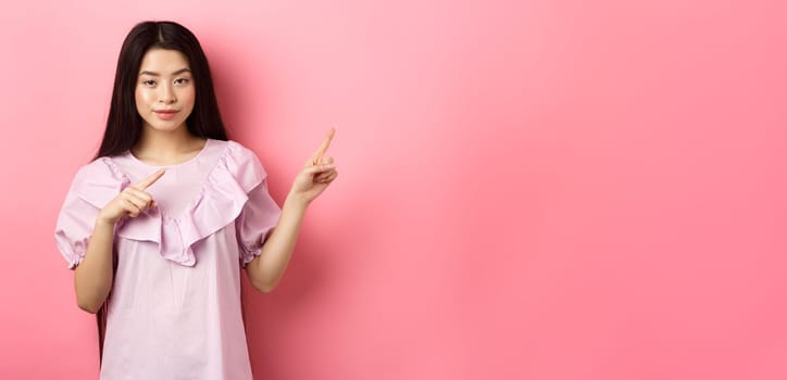 Cheeky asian girl showing interesting advertisement, pointing fingers right at logo and smiling cunning, hinting on good deal, pink background.