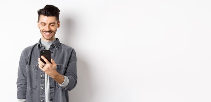 Handsome man reading message on phone and smiling. Guy holding smartphone and looking at screen, standing on white background.