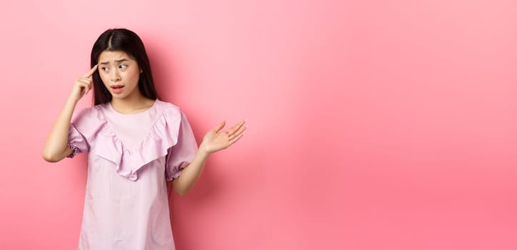 Are you stupid. Shocked asian woman pointing at head and complaining, scolding someone crazy or strange, look aside, standing against pink background.