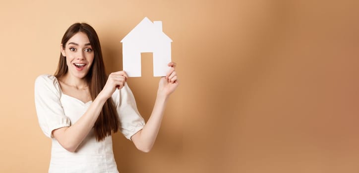 Real estate. Excited woman got new apartment, showing paper house cutout and smiling happy, standing on beige background.