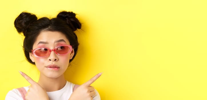 Summer fashion concept. Pensive asian girl making choice, pointing sideways and looking thoughtful, deciding what pick, standing in sunglasses on yellow background.