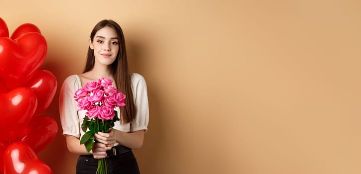 Beautiful girl holding bouquet of pink roses and smiling at camera, going on romantic date, standing near valentines heart balloons, beige background.