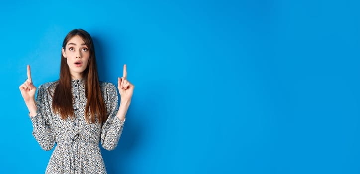 Surprised romantic girl in dress pointing, looking up with opened mouth and fascinated gaze, standing on blue background.