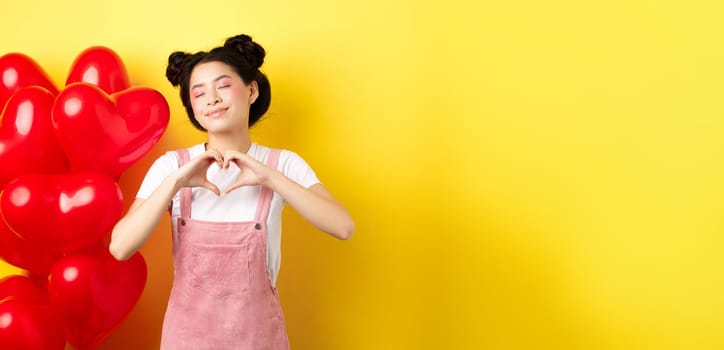 Valentines day concept. Cute asian girl dreaming of romance, close eyes and showing heart gesture, smiling happy, standing near red romantic balloons, yellow background.