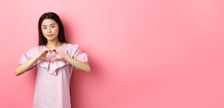 Valentines day concept. Beautiul japanese teen girl show heart gesture and smiling cute, express love and sympathy to lover, standing in romantic dress on pink background.