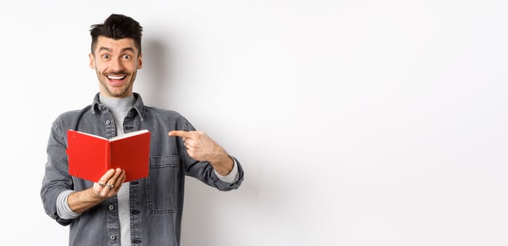 Happy smiling guy pointing at red planner, showing good journal or book, standing on white background.