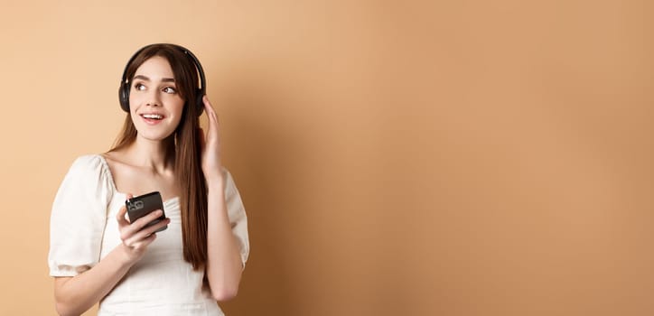 Attractive girl listening music in wireless headphones, holding mobile phone and looking aside with dreamy smile, beige background.