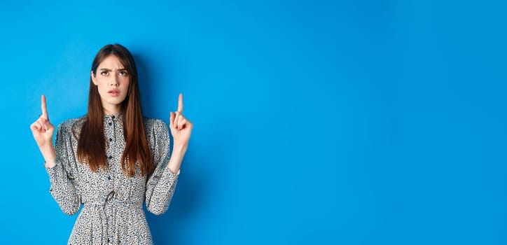 Confused attractive woman in dress frowning, pointing fingers up and looking puzzled, cant understand something, stare at strange thing, blue background.