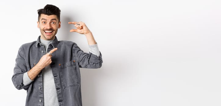 Excited man showing small size, pointing finger at little thing and smiling, standing against white background.