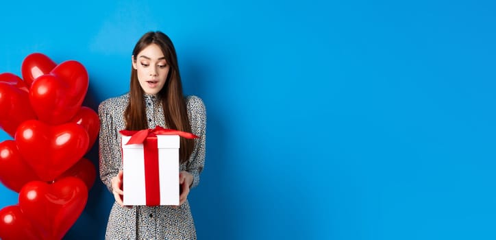 Valentines day. Image of beautiful girl looking surprised at gift box, receive present from lover, standing in dress on blue background.
