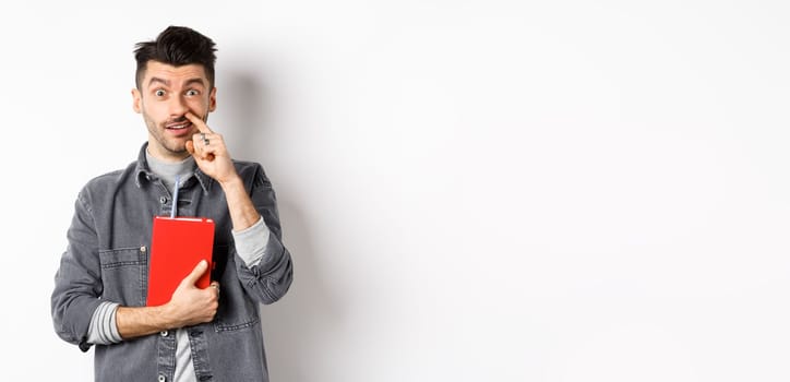 Young guy picking nose and holding book, look stupid at camera, standing against white background.