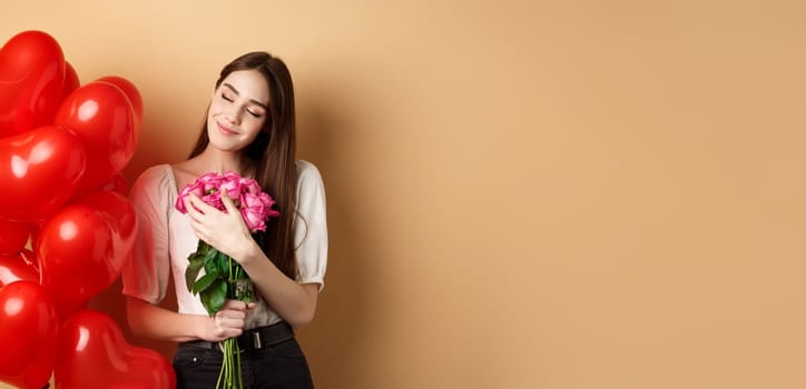 Sensual and romantic girl hugging bouquet of pink roses, smiling with eyes closed, thinking of lover, standing near Valentines day heart balloons, beige background.