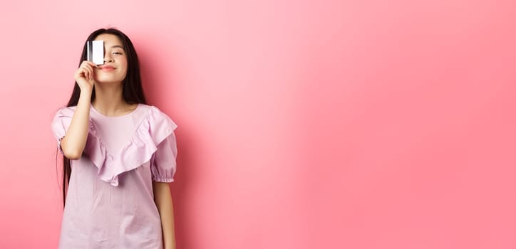 Shopping. Cute candid woman smiling, showing plastic credit card on face, standing in dress against pink background.