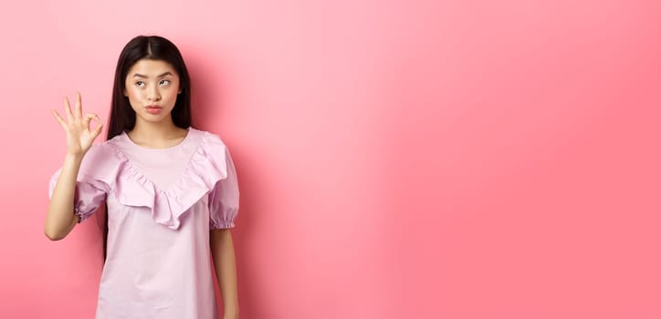 Sassy asian woman in dress show okay sign, looking aside, assure everything good, guarantee all OK, standing pleased on pink background.