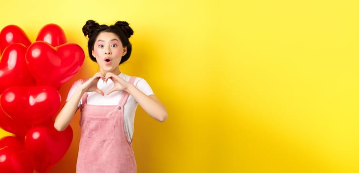 Surprised asian woman looking in awe, showing heart gesture, making love confession on valentines day, standing near romantic red balloons over yellow background.