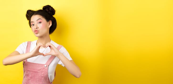 Silly japanese girl showing heart gesture and wishing happy Valentines day, pucker lips to kiss lover, standing on yellow background.