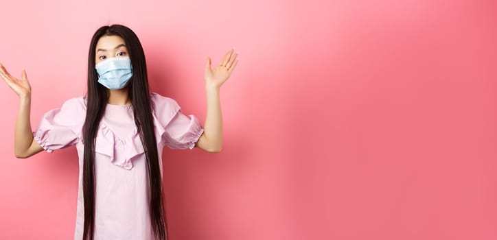 Coronavirus, quarantine and lifestyle concept. Clueless asian girl in face mask shrugging shoulders unware, know nothing, standing on pink background.