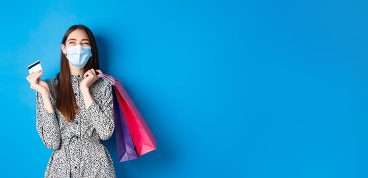 Covid-19, pandemic and lifestyle concept. Happy girl wear face mask on shopping, showing plastic credit card and laughing carefree, buying gifts, standing on blue background.