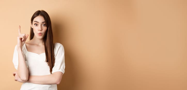 Creative girl pitching an idea, raising finger in eureka gesture, saying her plan, having a suggestion or solution, standing in dress on beige background.