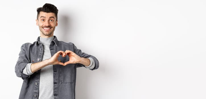 Smiling boyfriend showing heart with love, looking romantic and happy at lover, standing on white background. Valentines day and relationship concept.