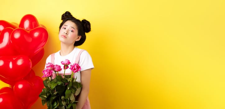 Happy Valentines concept. Sad and gloomy asian woman holding bouquet of roses and feeling upset and lonely on romantic lovers day, standing near red heart balloons, yellow background.