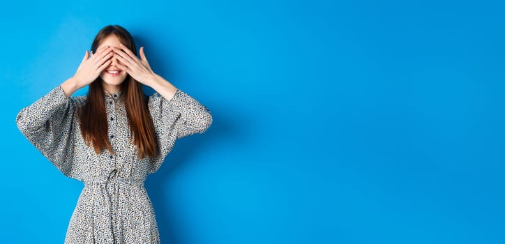 Happy smiling woman in dress cover eyes with hands, waiting for surprise, standing on blue background.