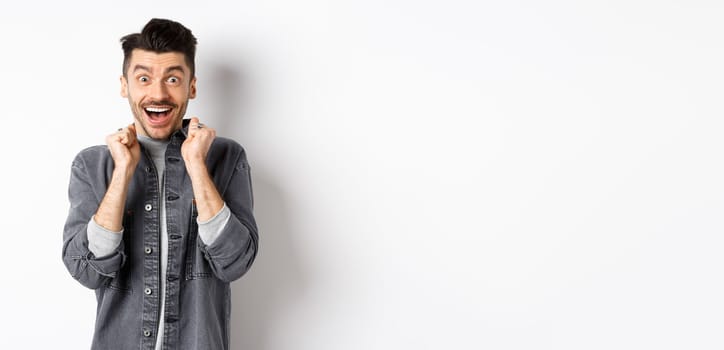 Excited young man jumping from happiness, smiling amazed and holding hands pressed to chest, standing on white background.