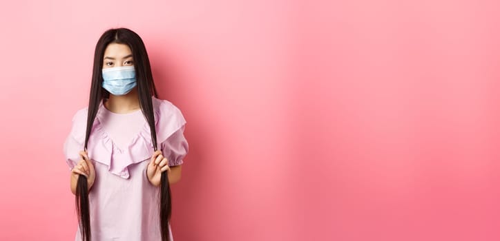 Healthy people and covid-19 pandemic concept. Sassy asian woman in medical mask playing with long hair, looking cool at camera, standing against pink background.
