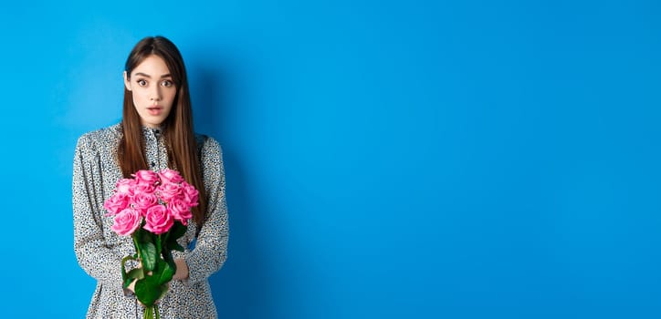 Valentines day concept. Surprised girlfriend receiving beautiful bouquet of flowers and looking with disbelief at camera, standing on blue background.