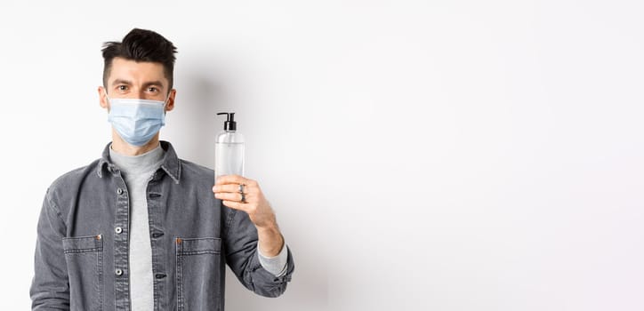 Healthy people and covid-19 concept. Young man in medical mask use preventive measures from coronavirus, showing bottle of hand sanitizer, standing against white background.