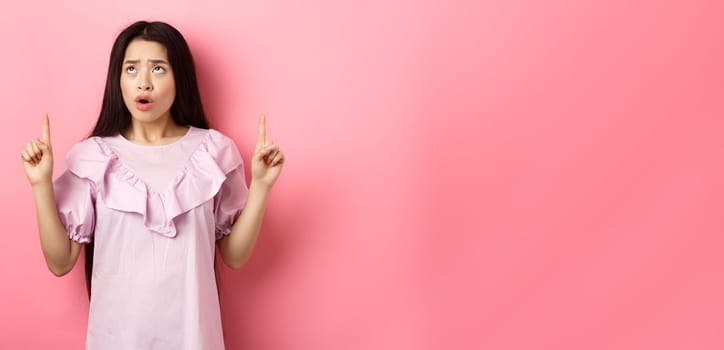 Concerned and sad asian girl frowning, looking and pointing fingers up at bad news, standing against pink background.