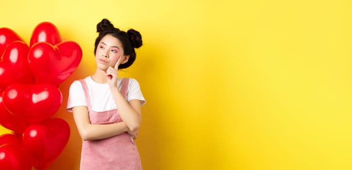 Valentines day concept. Pensive asian woman looking left, thinking about romantic date, standing near red hearts balloons on yellow background.