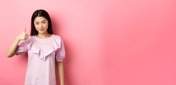 Smiling asian girl show thumb up in approval, praise good choice, like and recommend product, standing on pink background.