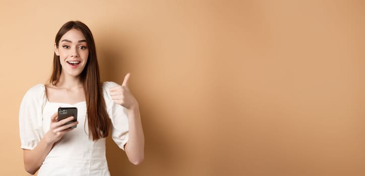 Online shopping concept. Happy young woman showing thumb up and using cellphone, smiling pleased, standing on beige background.