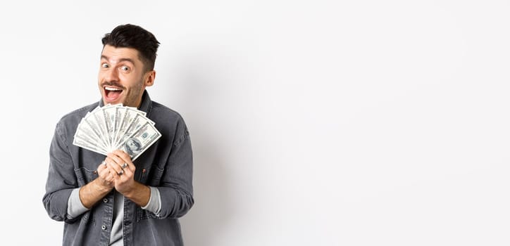 Excited young man smiling and showing dollar bills, making money, standing against white background.