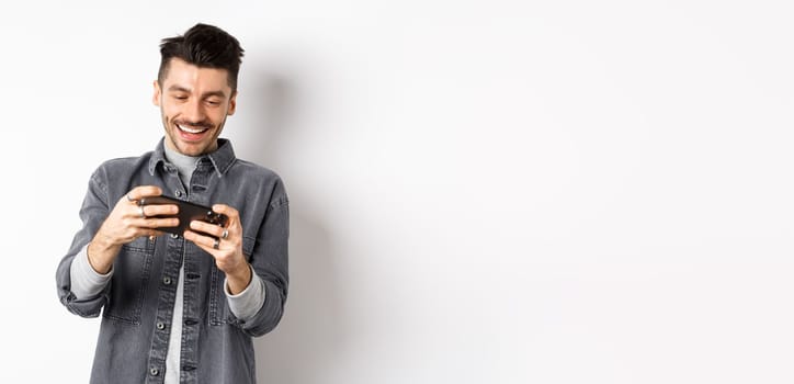 Handsome young man playing mobile video game and smiling at screen, standing against white background.