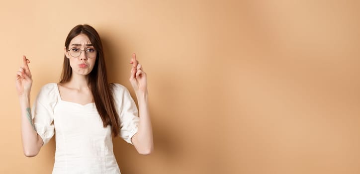 Cute hopeful girl making wish, pucker lips and begging, say please, standing on beige background.