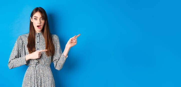Wow look that way. Excited young woman with long hair and trendy dress, drop jaw and pointing right at logo, showing awesome news, blue background.