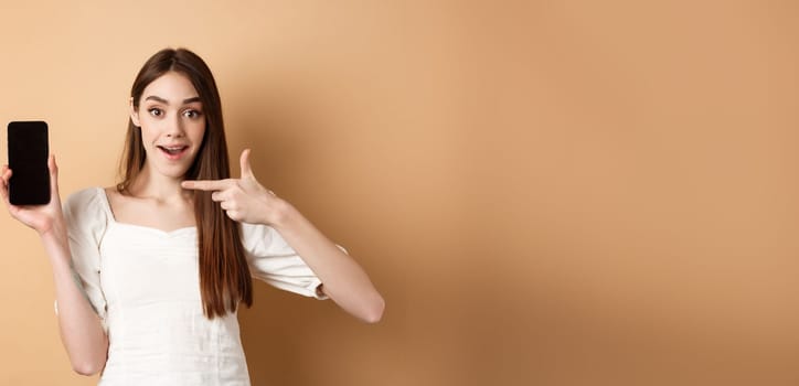 Cheerful woman pointing finger at empty phone screen, looking excited, standing on beige background.